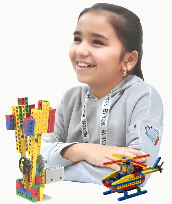 A young girl with dark hair tied back smiles while sitting at a white table, looking at a colorful toy model she built with interlocking blocks. The model features a yellow tower with red, blue, and green accents, alongside a small yellow and blue helicopter.