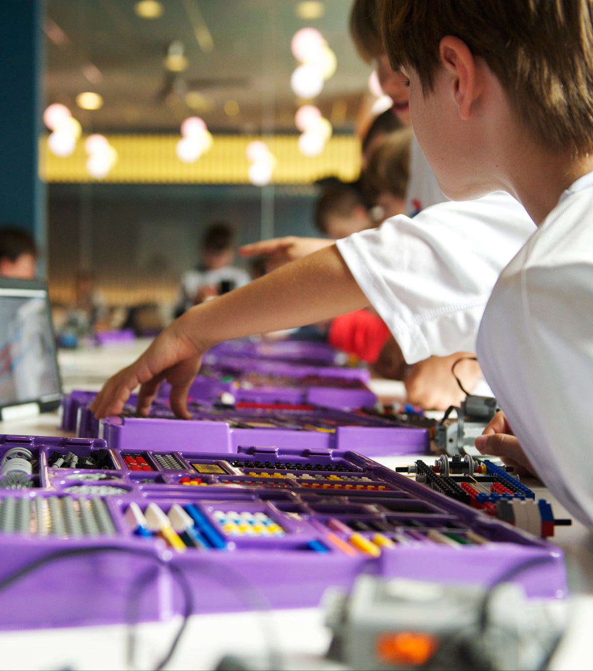A person in a white shirt works at a table with a purple robotics kit in a bright indoor lab or classroom setting.